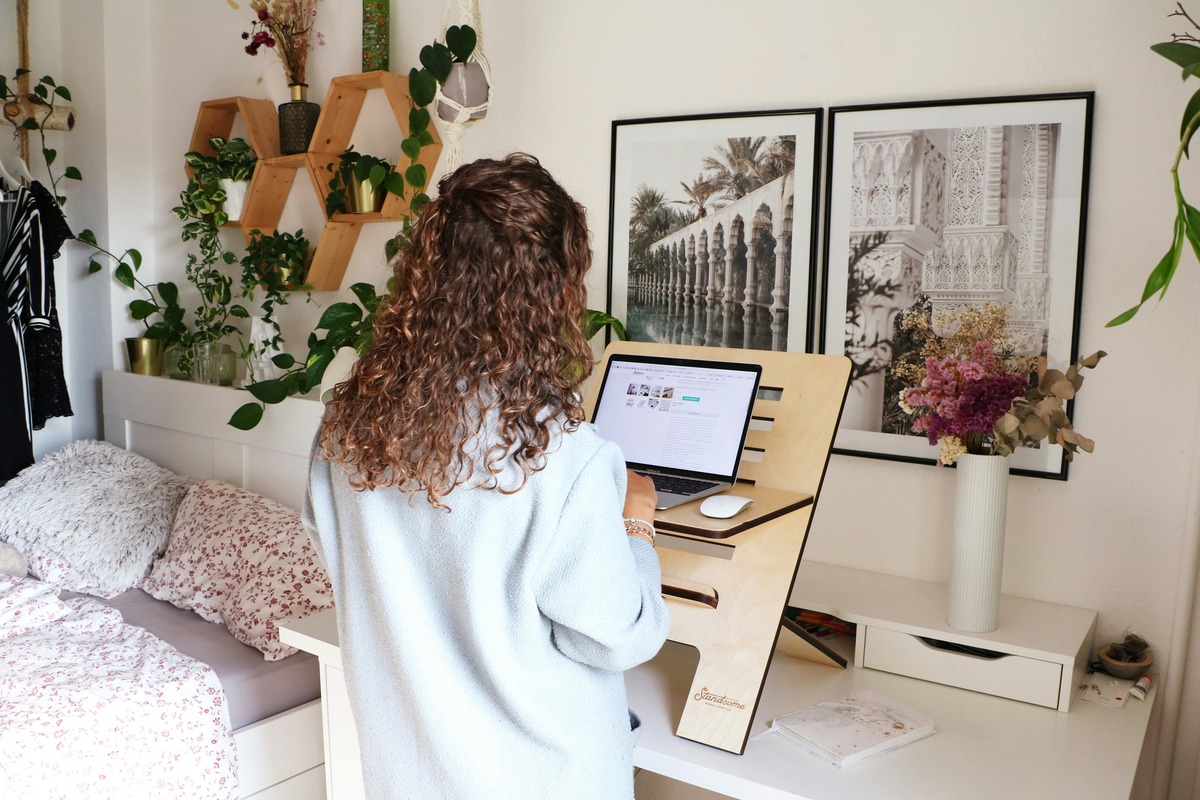 Woman with curly hair using her laptop at a standing desk in her bright, organized bedroom workspace